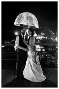 A black and white photo of a bride and groom under a clear umbrella in the night rain with backlighting