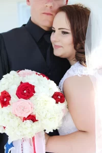 Picture of a bride leaning into her groom and holding her bouquet of white and red flowers