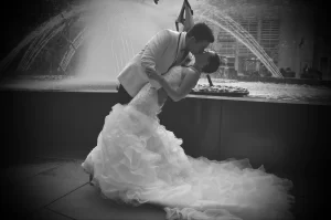 A black and white photo of a bride and groom kissing in front of a fountain