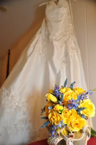 A bridal gown hanging up and her bouquet of yellow roses in front of it